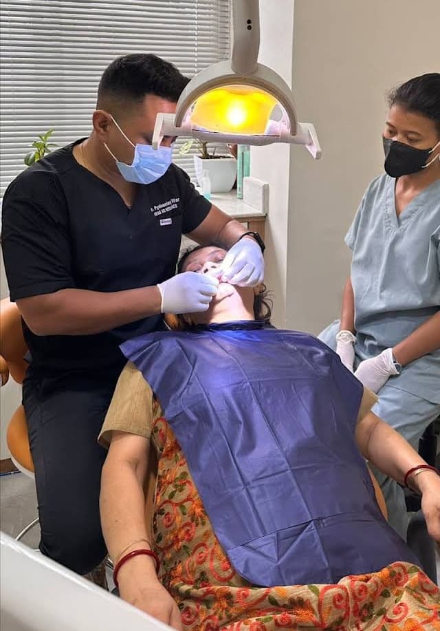 Dentist in black scrubs examines a patient's mouth while an assistant watches in a clinic.