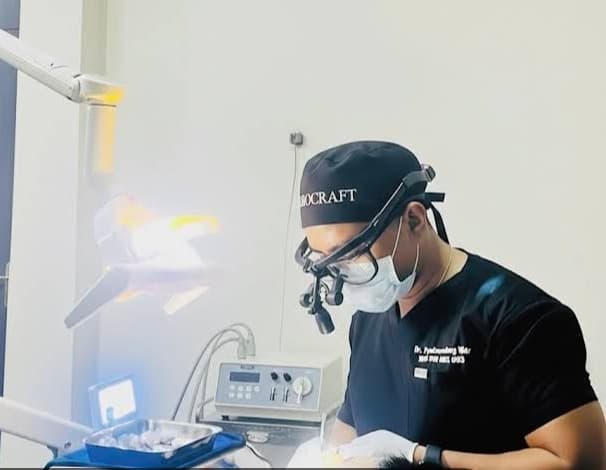 Dentist in black scrubs and surgical loupes performs a procedure under bright clinical lights.