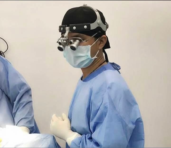 Surgeon wearing blue scrubs, a face mask, and magnifying loupes during a medical procedure.