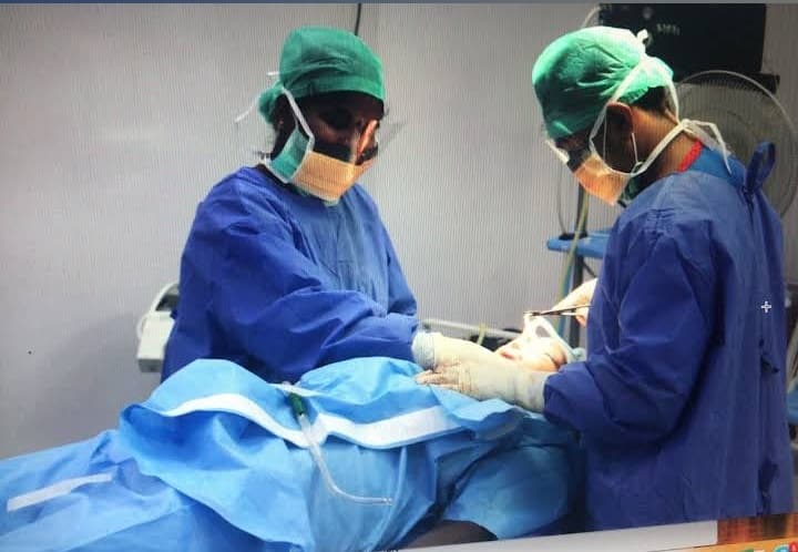 Two surgeons in blue scrubs and green caps perform surgery on a draped patient.