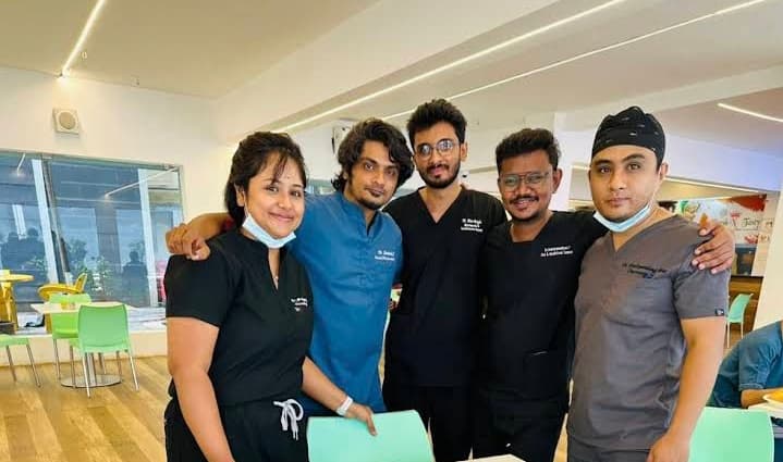 Five healthcare professionals in medical scrubs posing together in a brightly lit indoor space.